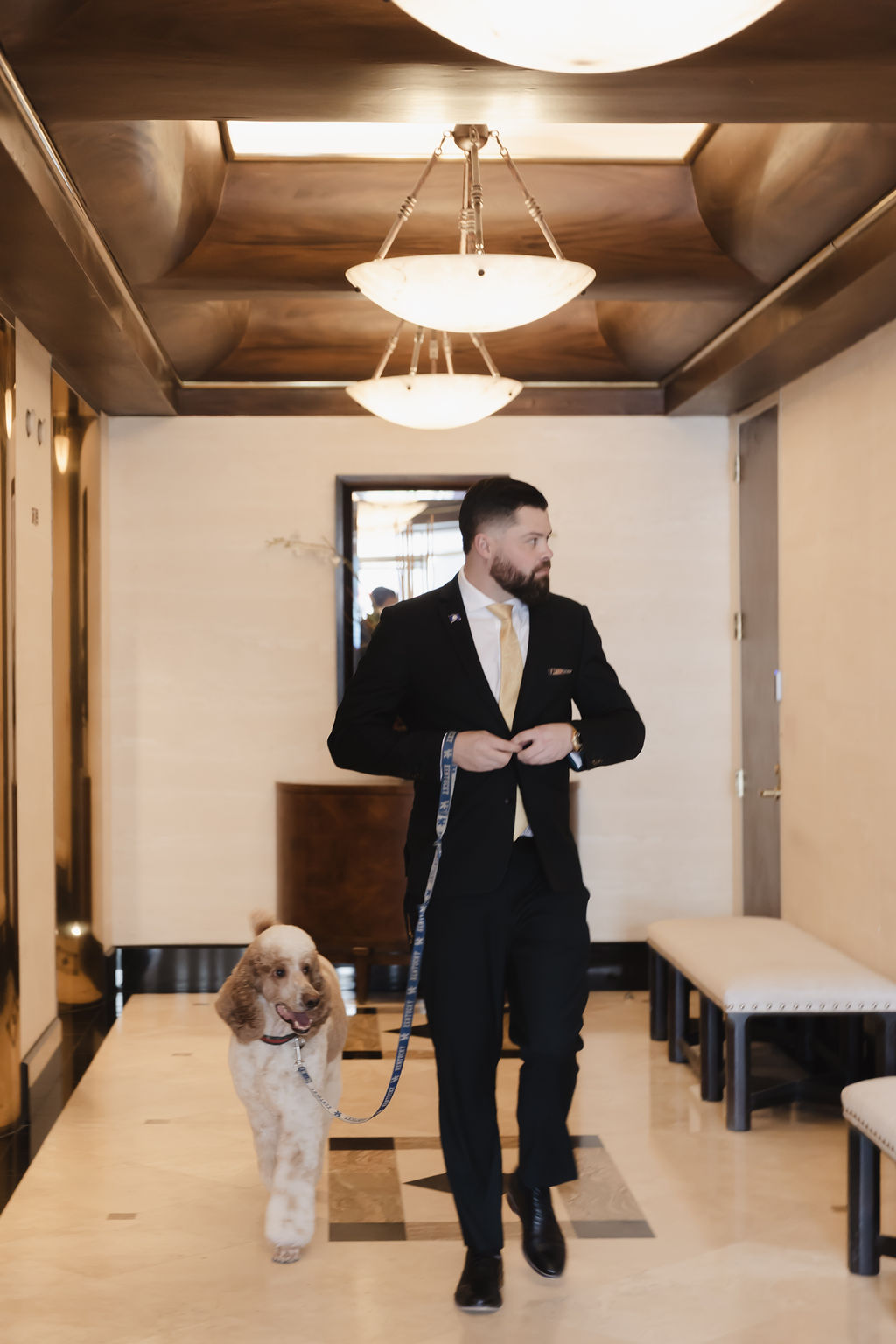 Bart Denham in a black suit and gold tie walking a fluffy tan dog on a leash through a brightly lit hotel hallway.