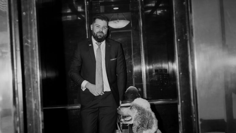 Bart Denham in a suit and tie standing in an elevator with a poodle.
